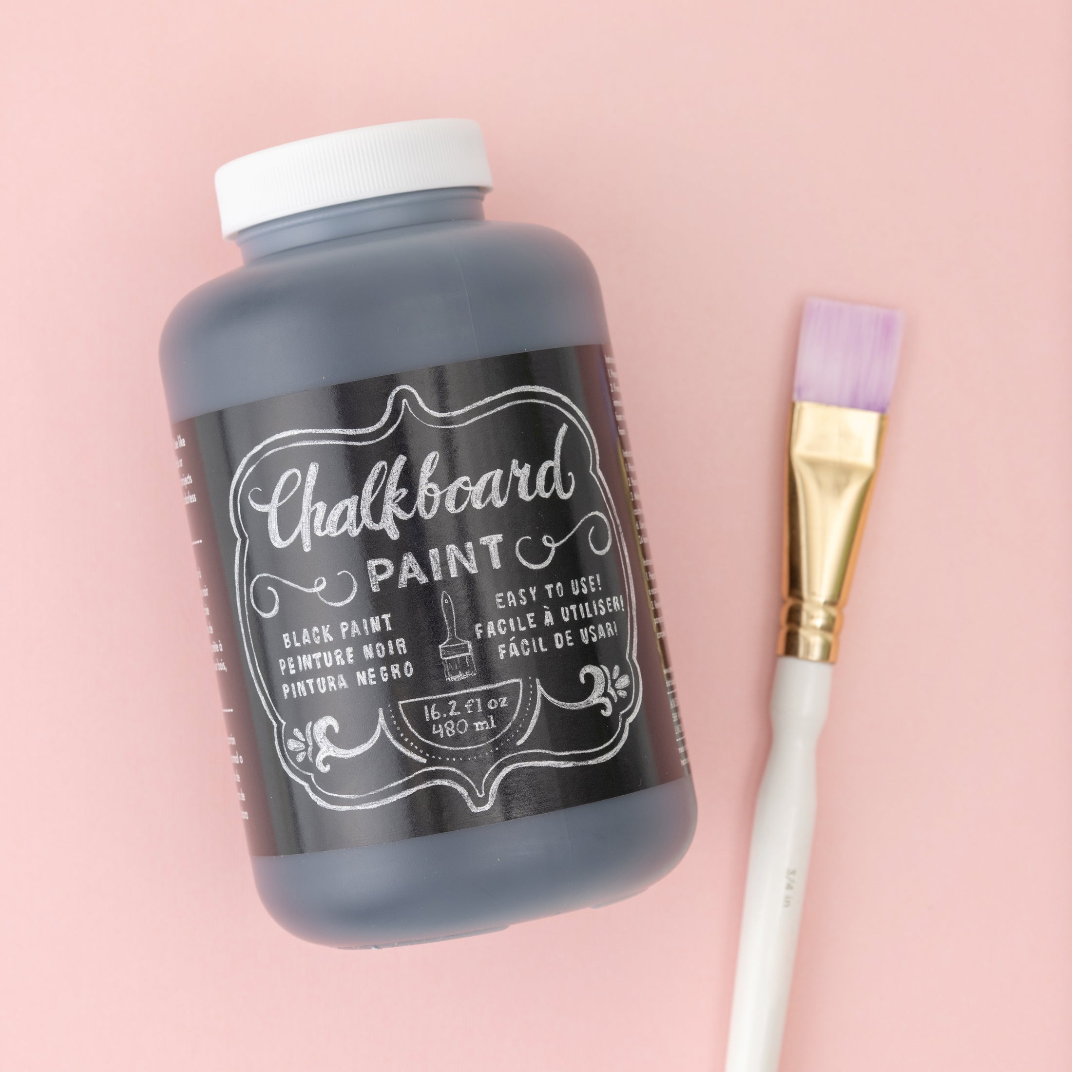 Lifestyle photo of American Crafts 16 oz Chalkboard Black Paint on pink back round with paint brush next to it