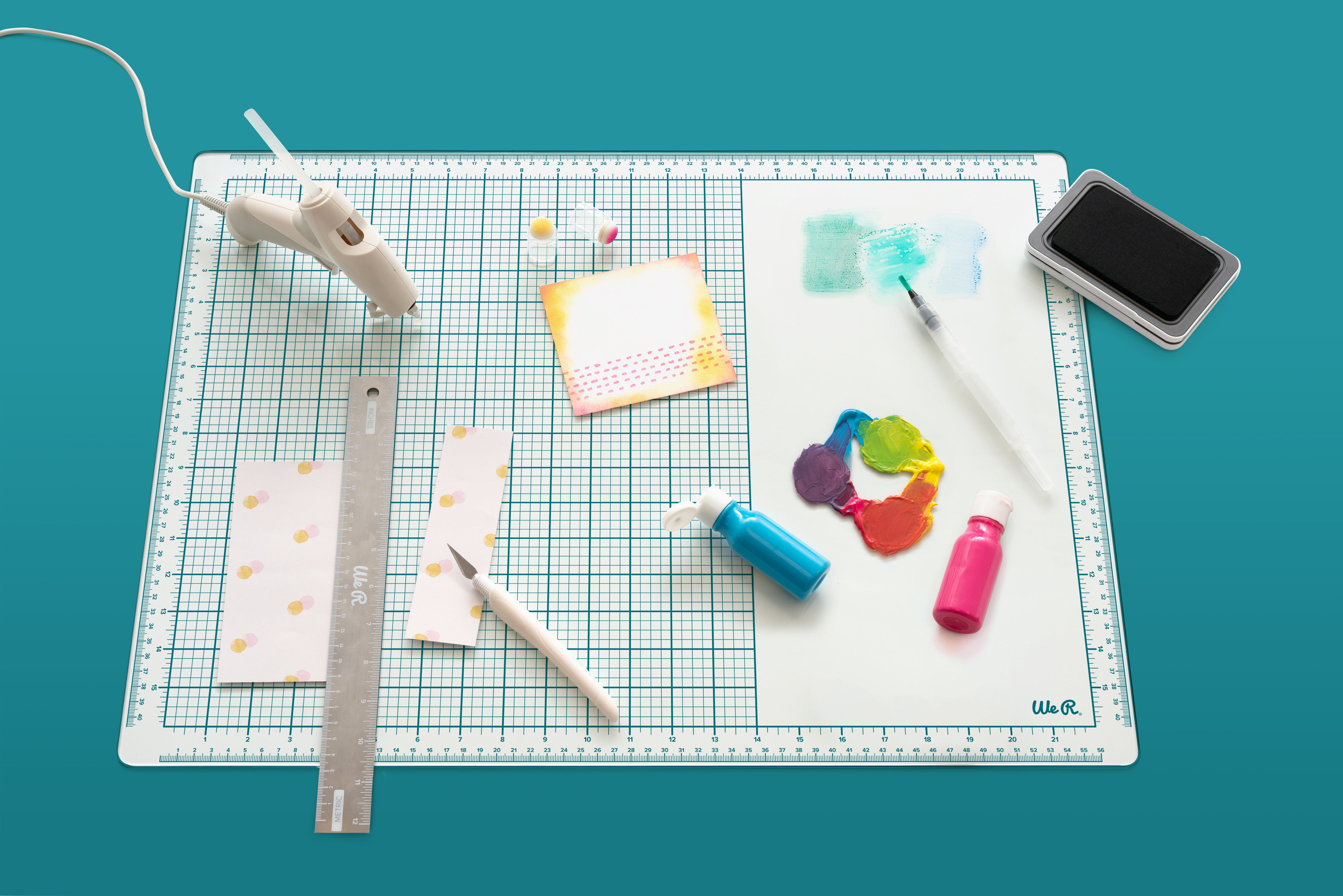 Picture of some crafts like paint, glue gun, ruler, and other items on the We R Makers Mixed Media 16x22 Glass Mat.